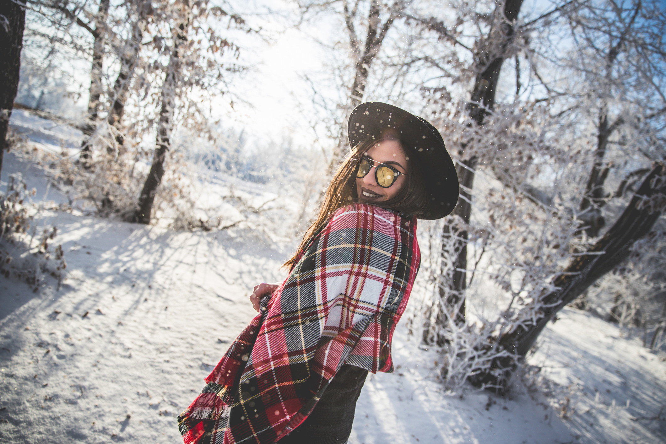 A woman looking over her shoulder as she walks through the snow while wearing a plaid blanket scarf, sunglasses, and a wide-brimmed hat