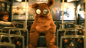 A furry bear-like mascot with bug-eyes sits on a bus