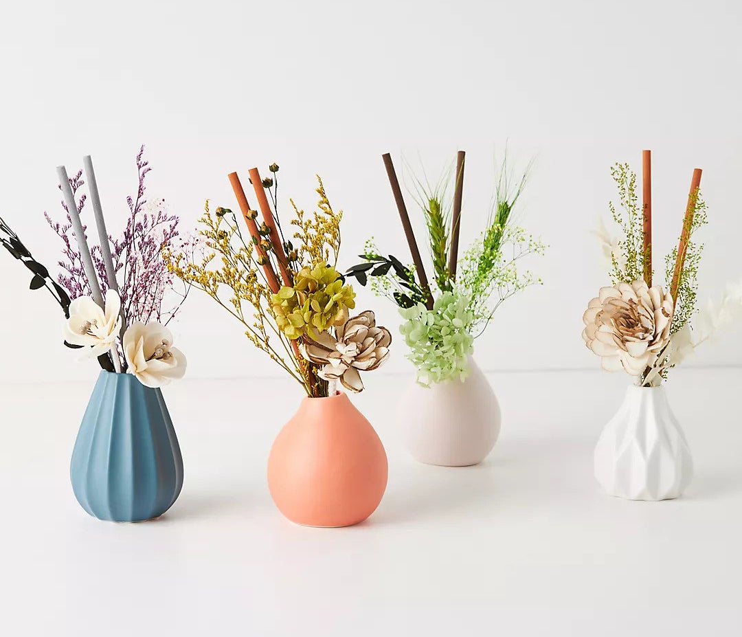 Four ceramic diffusers with dried florals and diffuser reeds in four different colors: blue, pink, lavender, and white