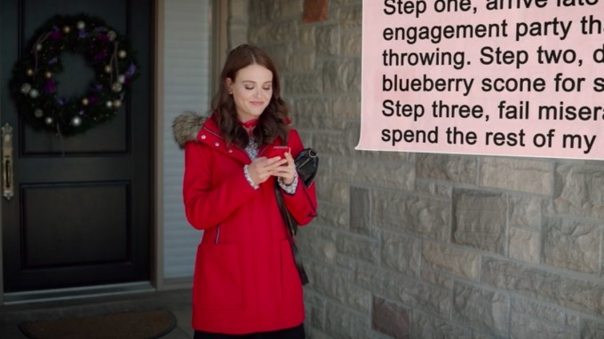 A woman with brown hair stands outside a house, texting. She&#x27;s wearing a red coat and smiling.