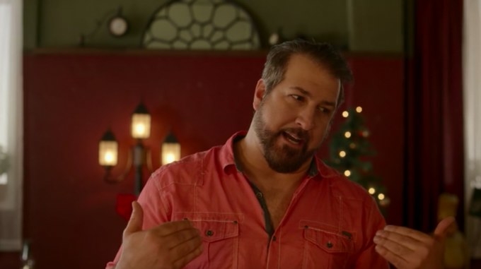 A man in a pink shirt stands in front of a Christmas tree, gesturing at himself.