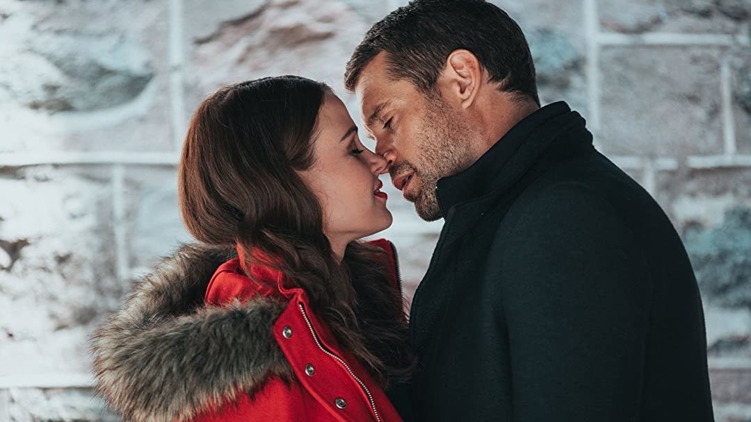 A couple stand outside a building, about to kiss. She&#x27;s wearing a red coat, he&#x27;s wearing a black coat.