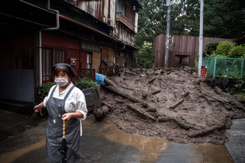 A woman in a mask and apron holds an umbrella with a mudslide behind her