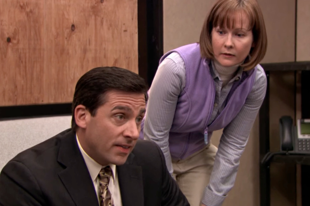 Steve Carrell is on his knees performing CPR on an unseen patient dummy while a woman in purple observes him in a still from The Office