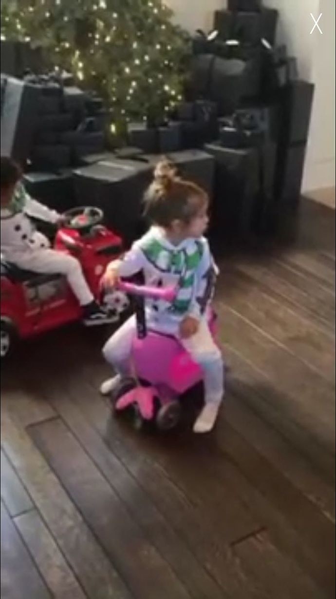Children riding toy scooters and firetrucks around the room filled with gifts
