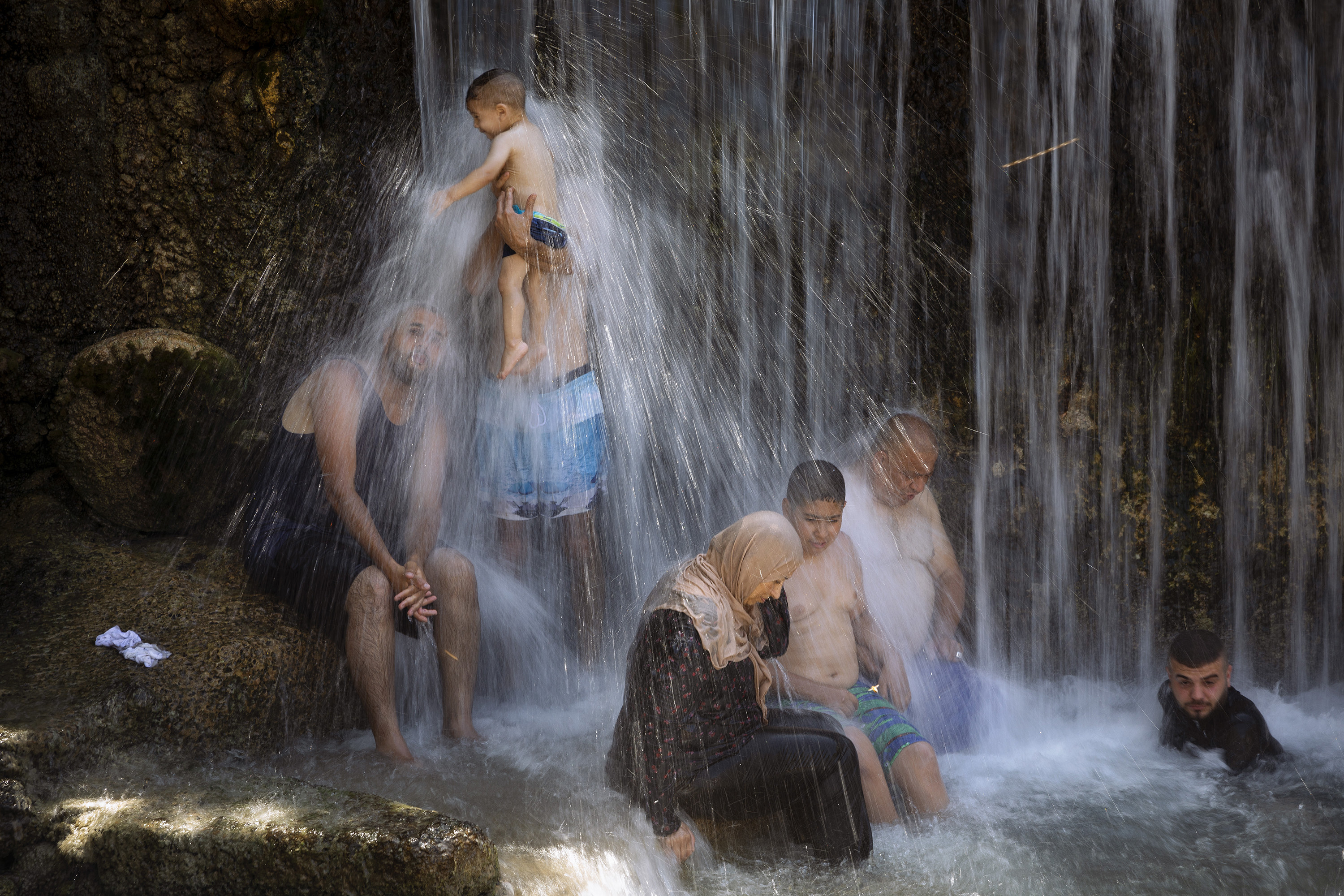 A man holds a baby under the waterfall as other people sit nearby