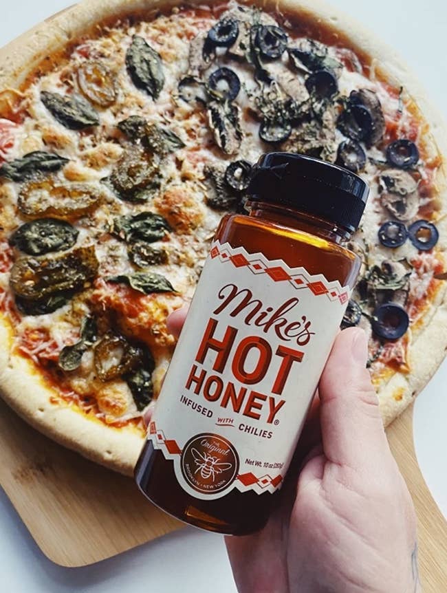 a hand holding a bottle of the honey in front of a pizza drizzled with it