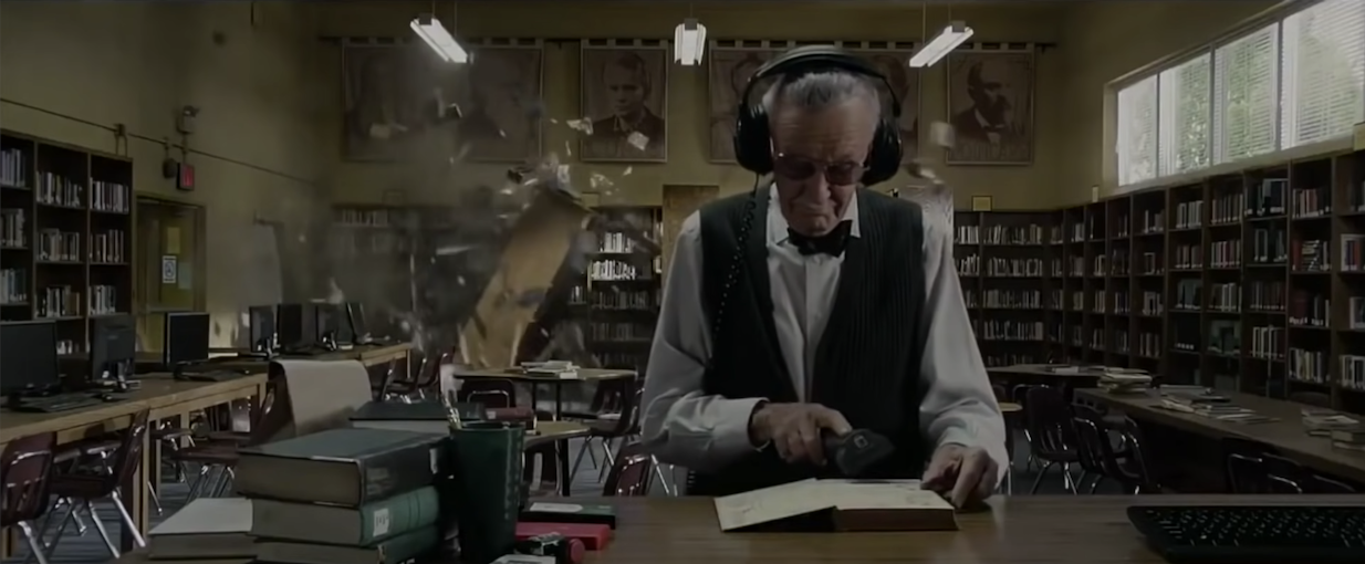 Stan Lee wearing headphones in the library, unaware of the Spidey battle behind him