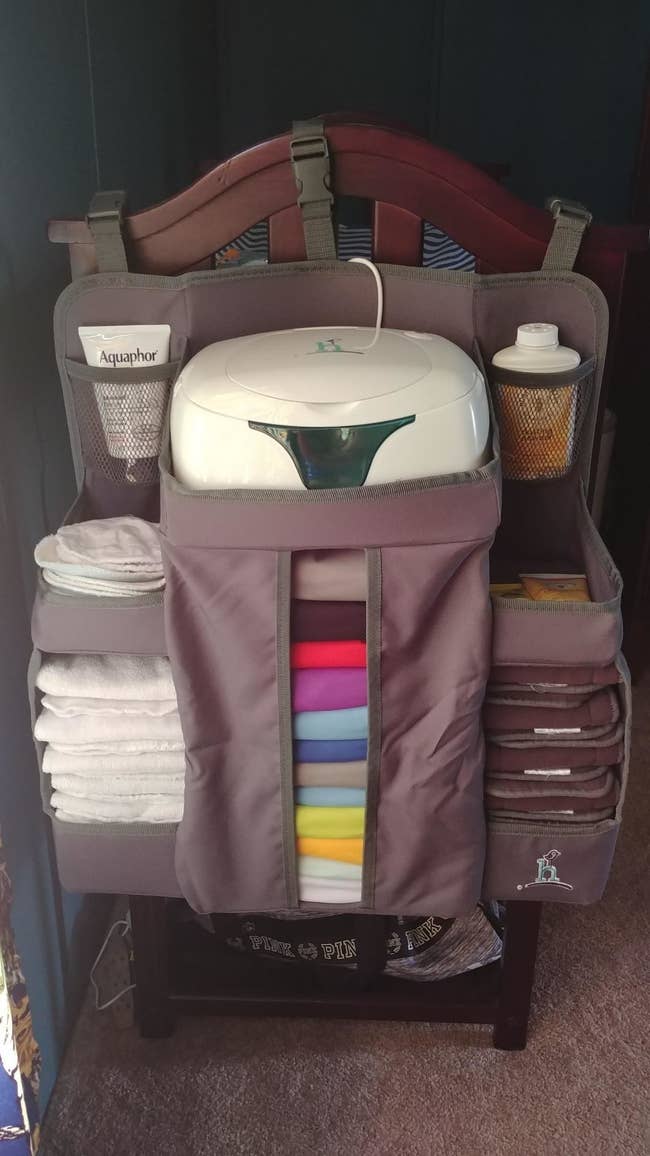 reviewer's large gray organizer hanging on edge of crib with slots for diapers, wipes, powder, lotion, and more