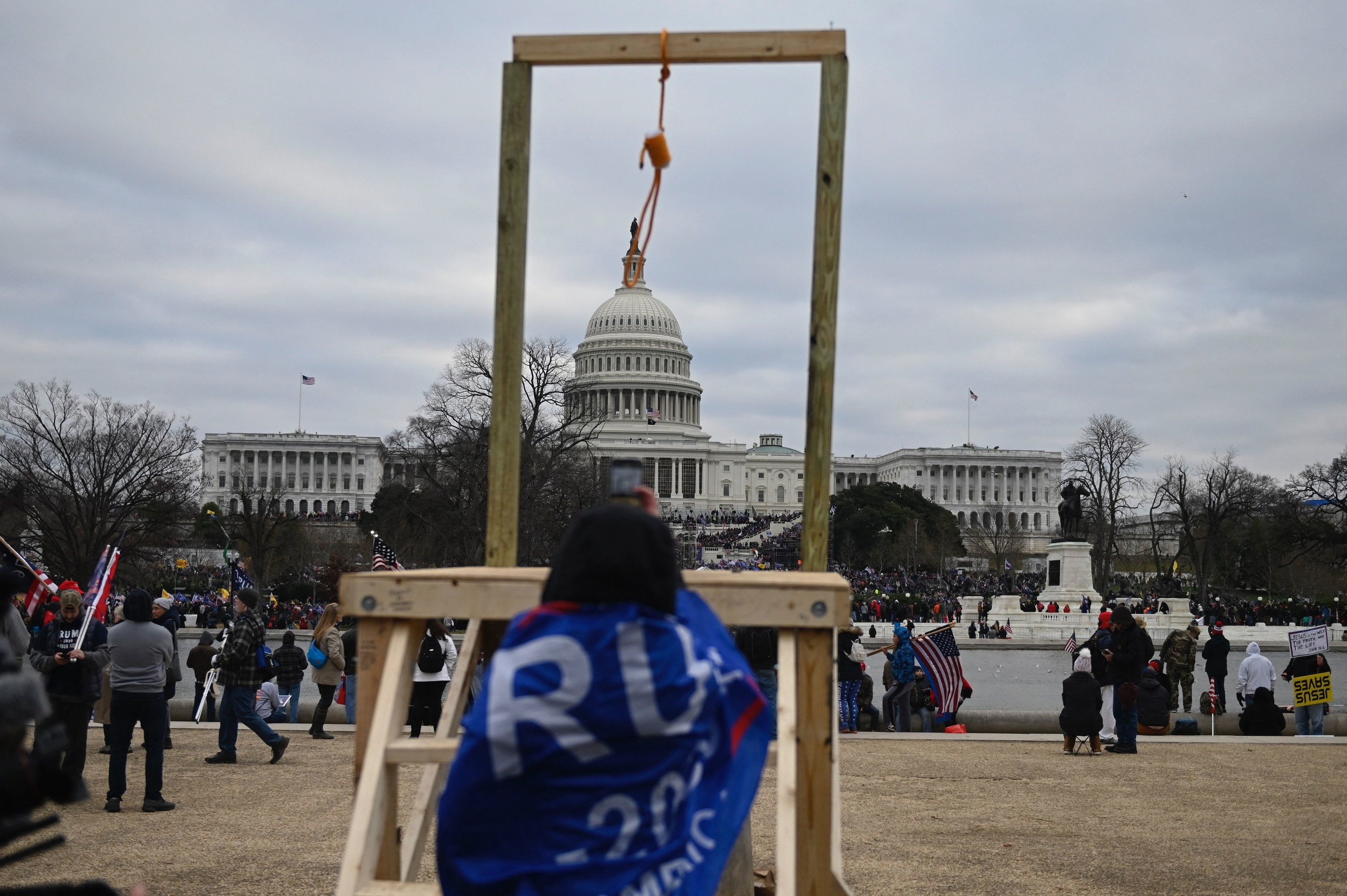 A woman wearing a Trump 2020 flag as a cape stands in front of a noose that was erected in front of the Capitol building
