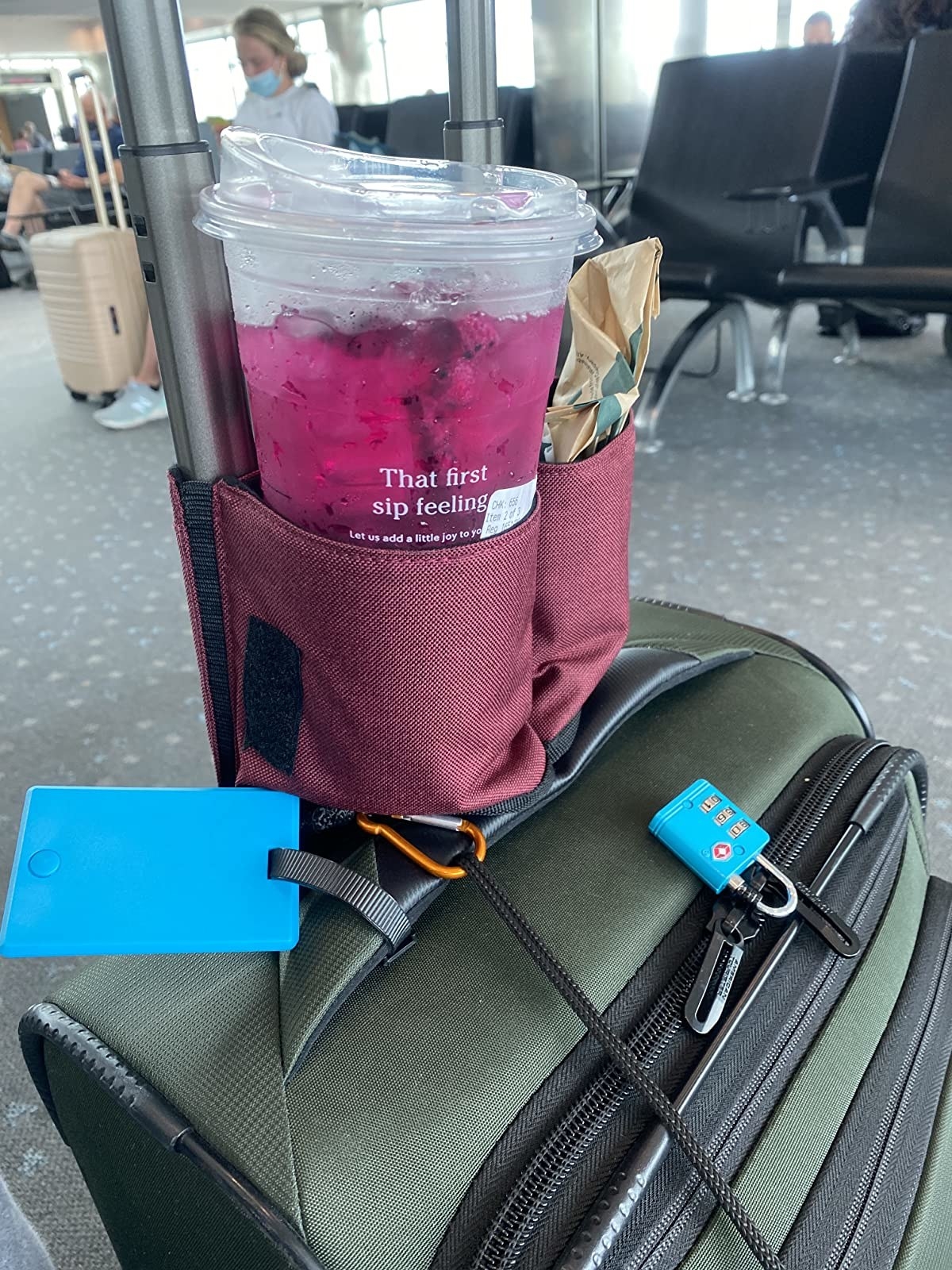 a travel cup holder attached to the handle of someone&#x27;s luggage, holding a cup from starbucks in one side and a bag of food in the other