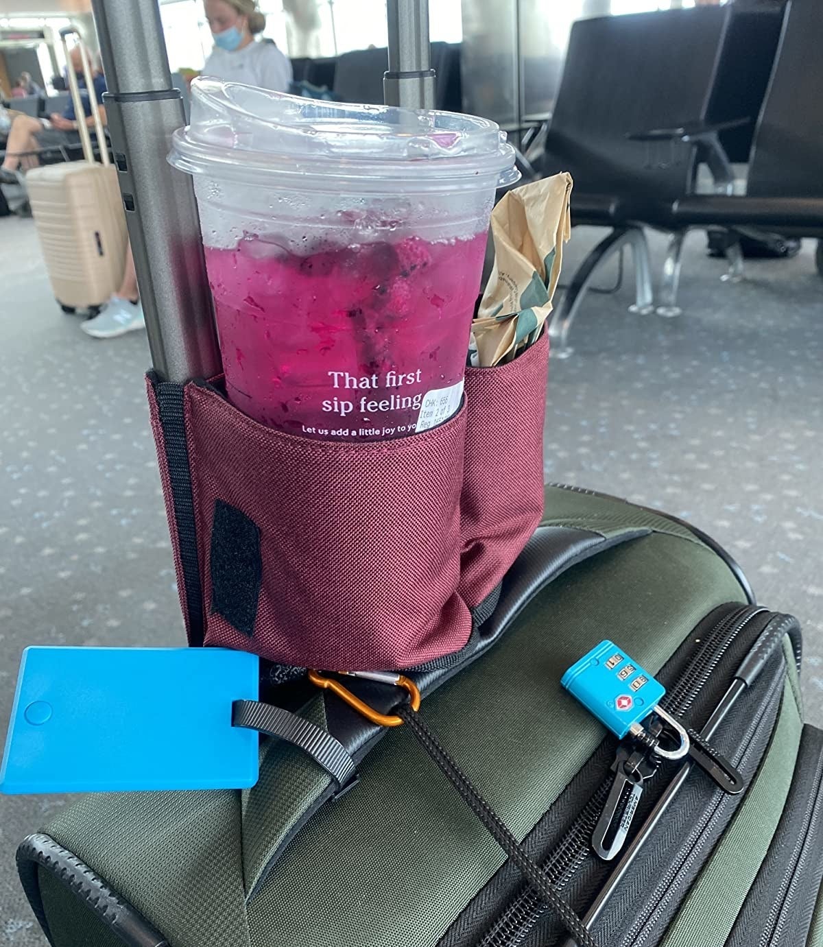 a travel cup holder attached to the handle of someone's luggage, holding a cup from starbucks in one side and a bag of food in the other