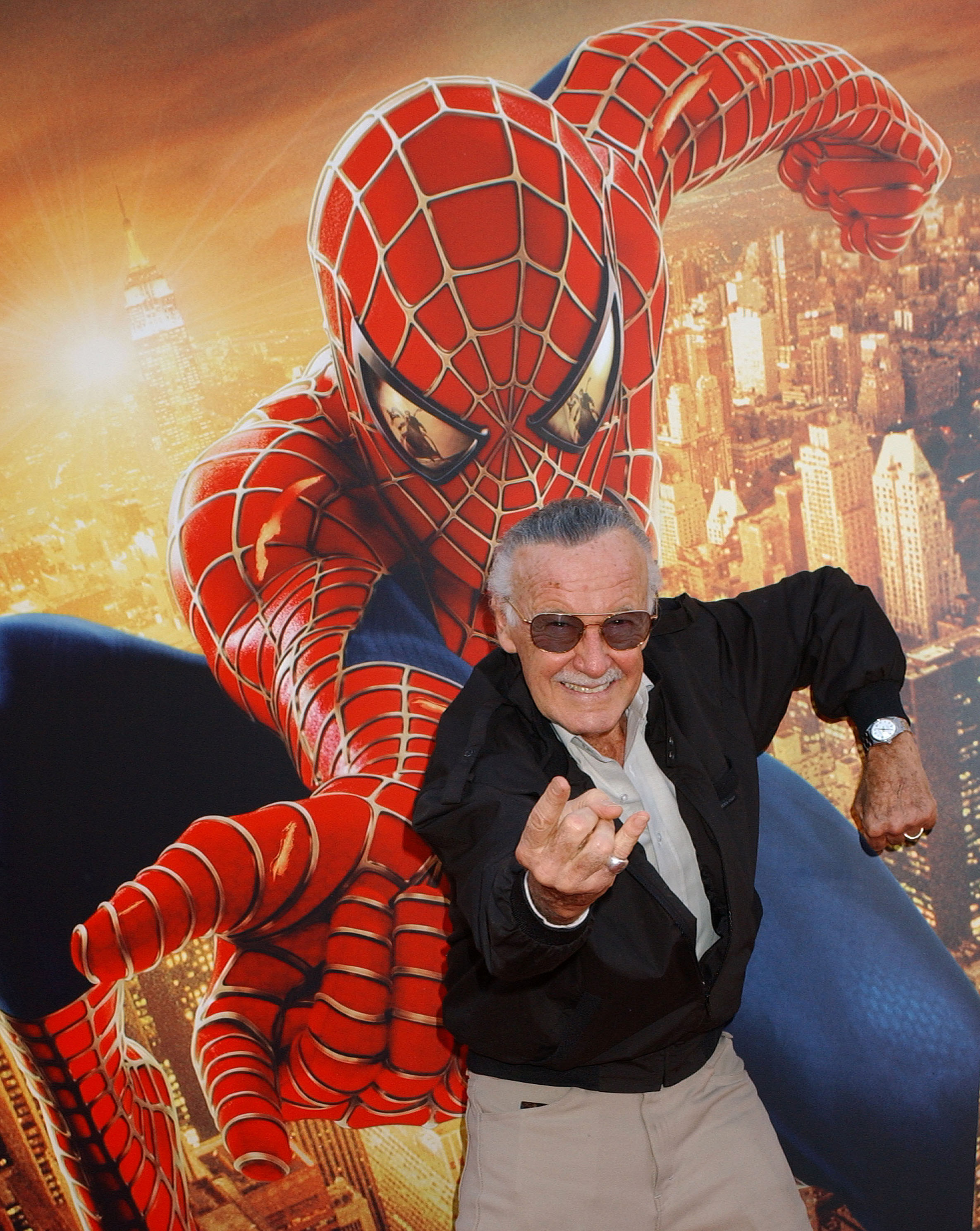 Stan Lee posing as if he&#x27;s shooting a spider&#x27;s web from his wrist in front of a Spider-Man poster