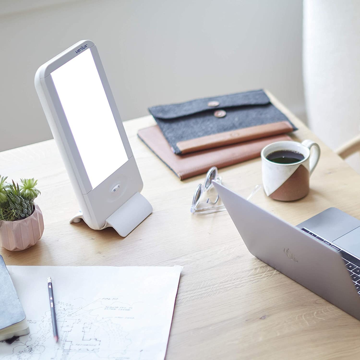 A light therapy lamp sits on a wooden desk, amidst various office paraphernaelia (a sketch, a mug of coffee, an open laptop, a laptop sleeve, a potted plant, a pair of sunglasses)