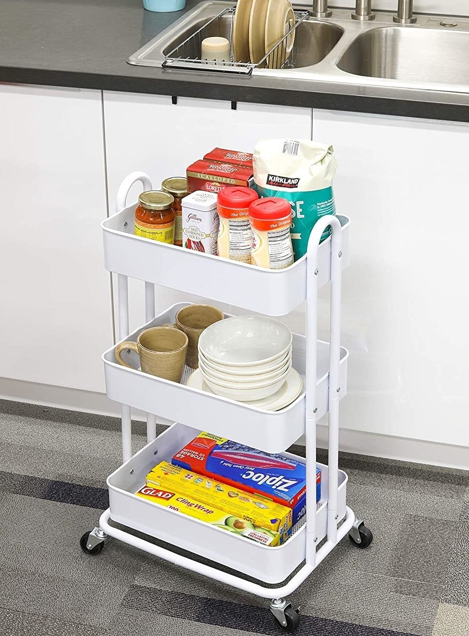 A metal rolling utility cart with spices in one shelf, plates and bowls on the second shelf, and plastic food wrap boxes on the lowest shelf