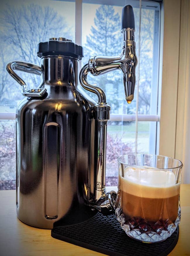 Stainless steel cold brew maker with curved spout next to a fresh cup of coffee
