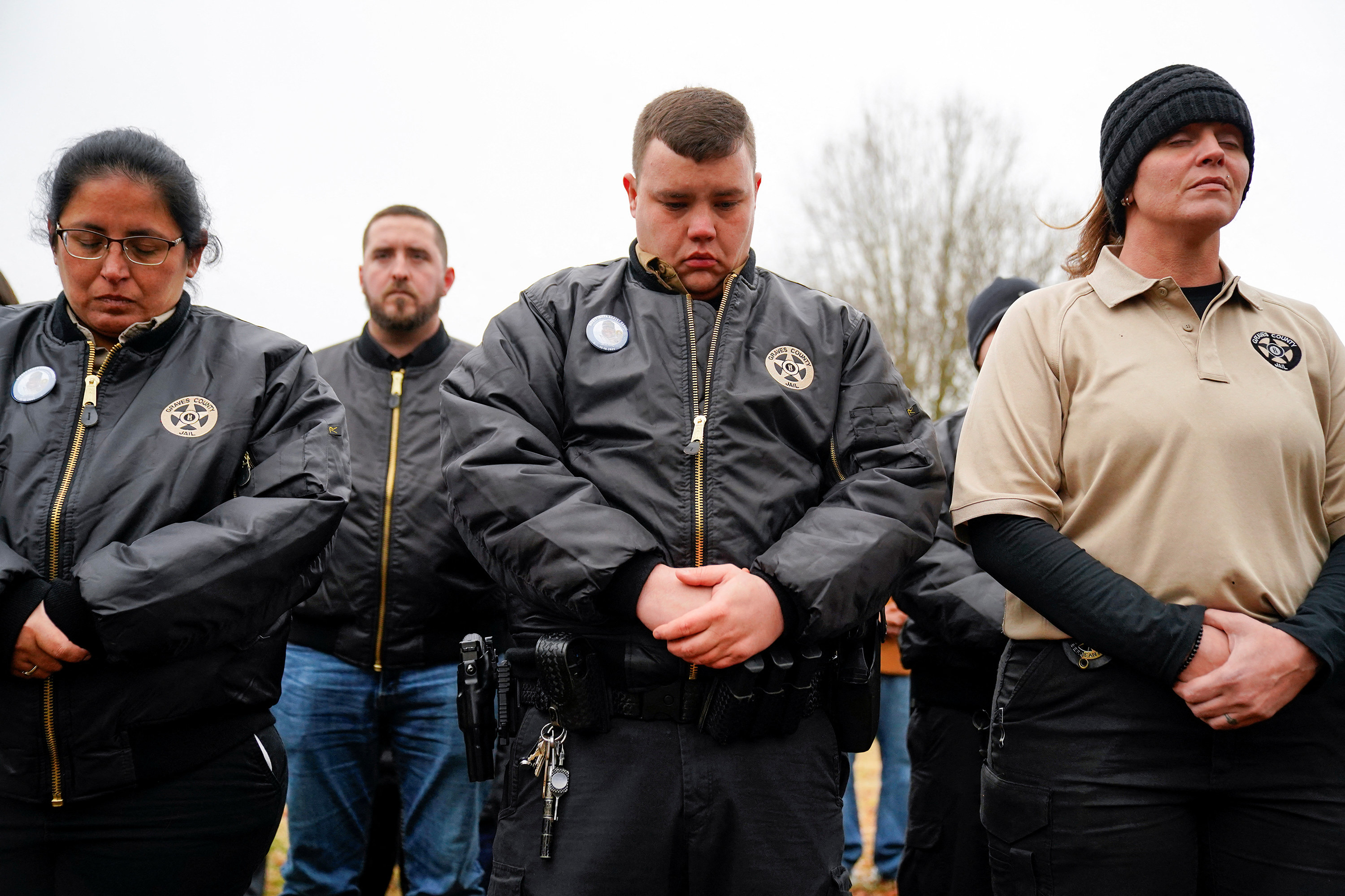 Four law enforcement officers stand with their hands clasped, looking down, with a tree in the background 