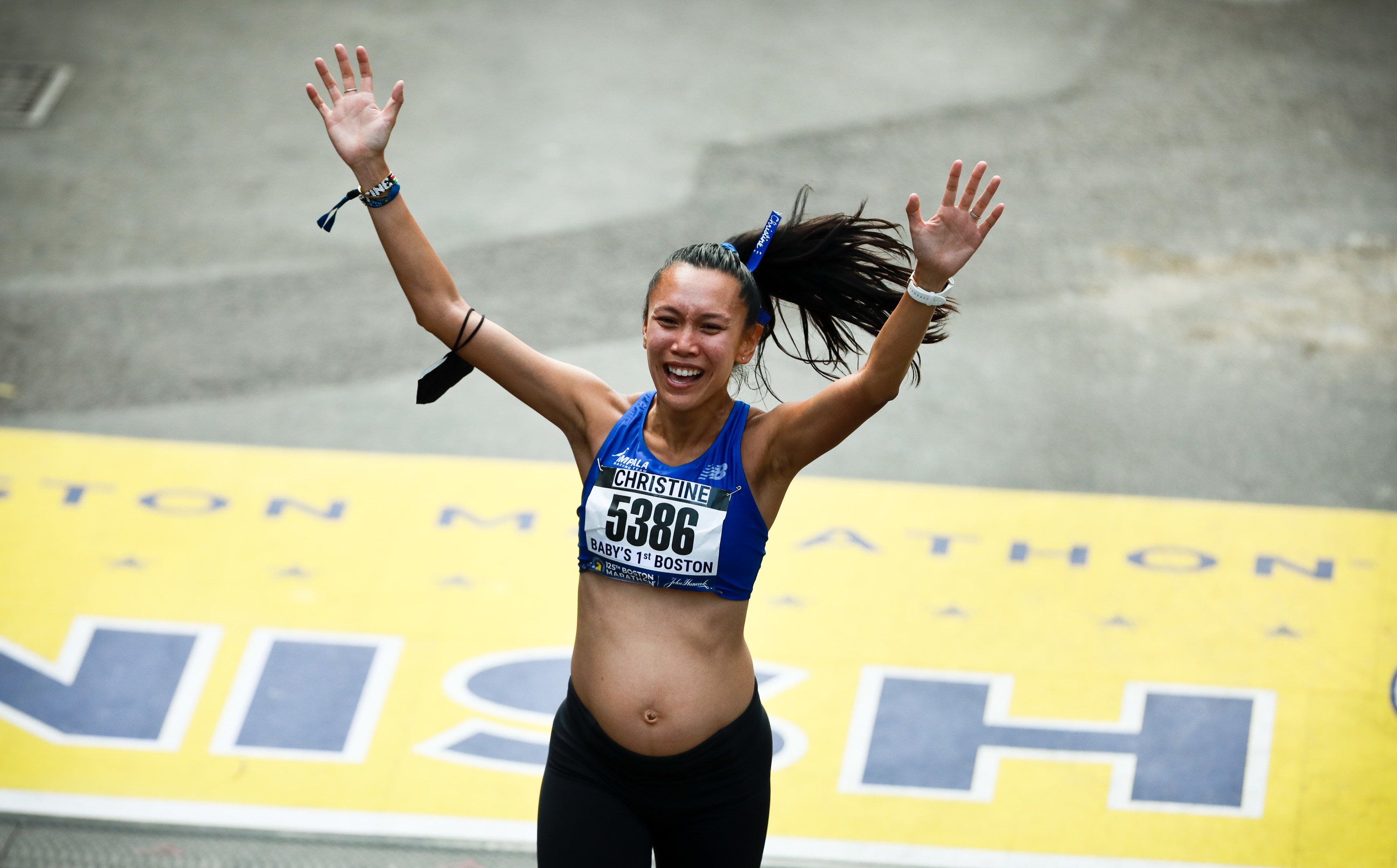 A smiling pregnant woman with her hands up crosses the finish line of a race