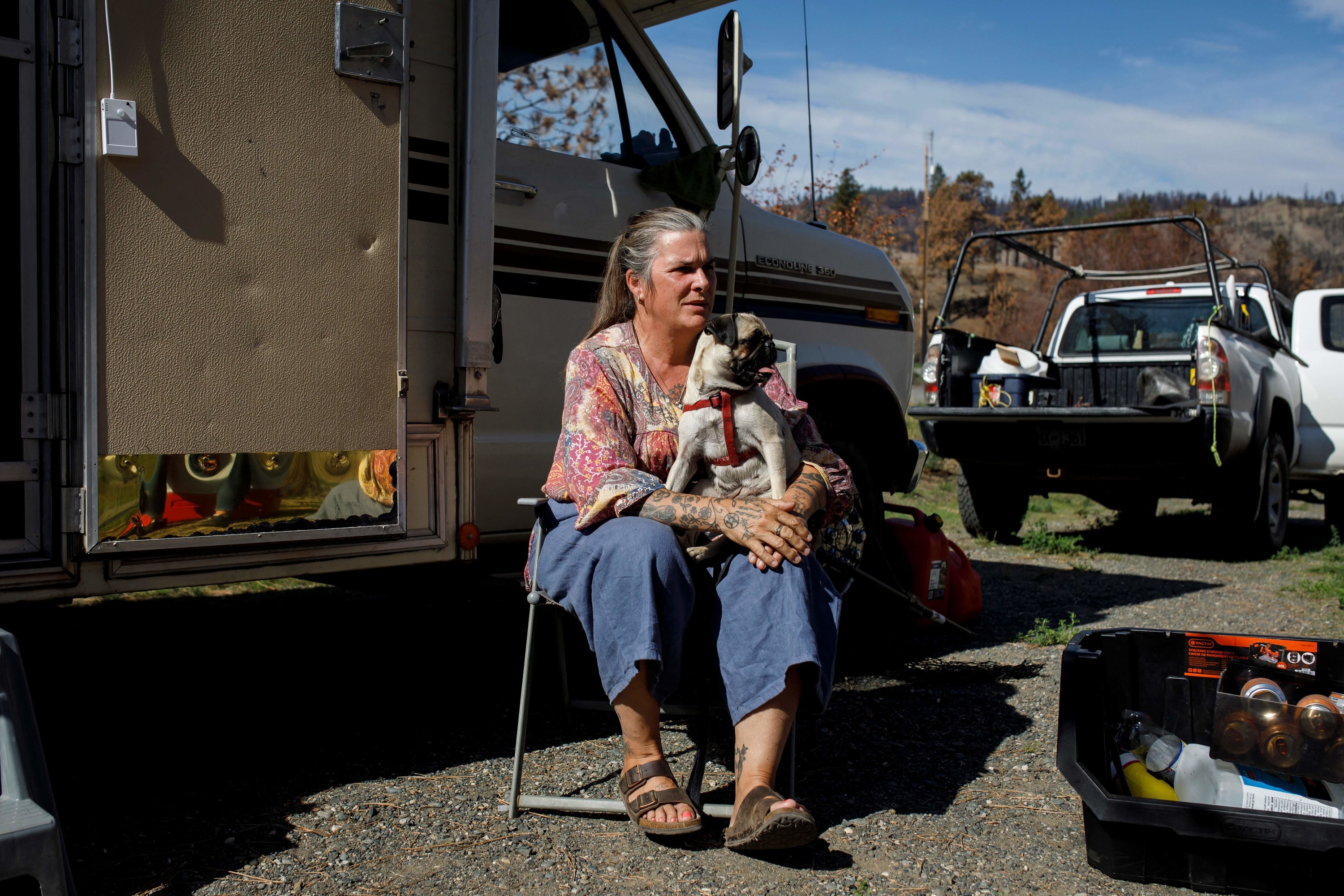 A woman sits outside a trailer holding her pug and wearing Birkenstocks with a truck behind her 
