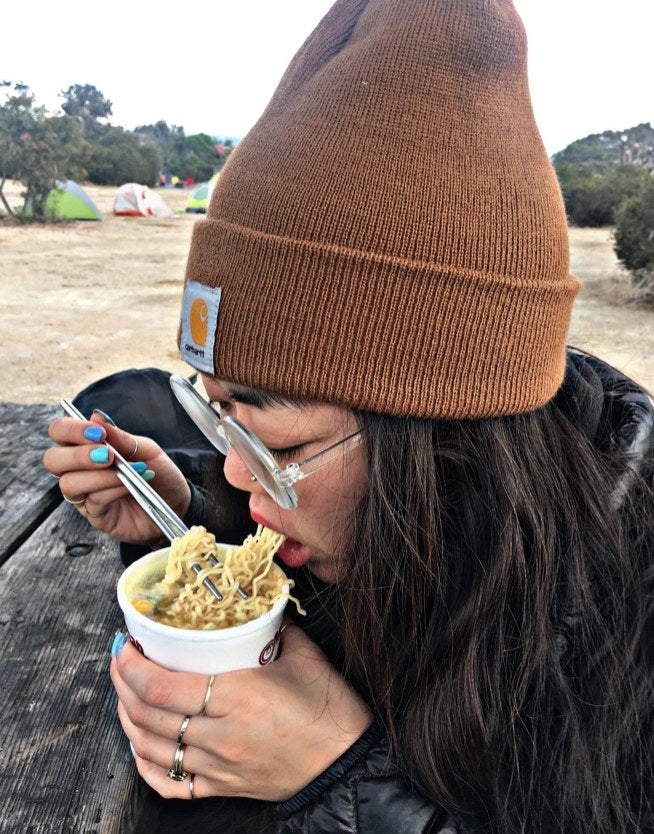 A reviewer wearing a brown hat with black jacket while eating noodles
