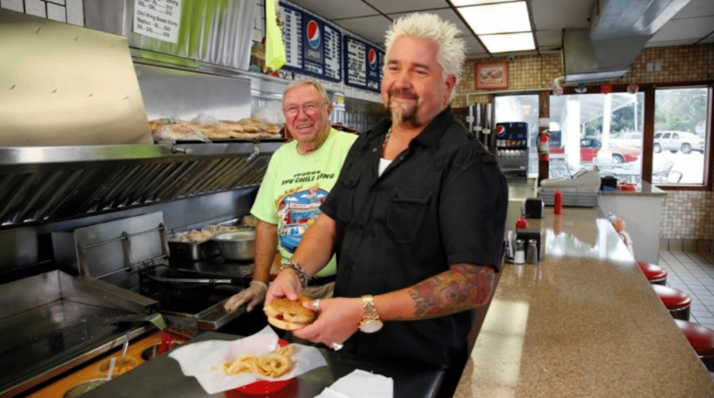 Guy Fieri stands with a burger in a diner