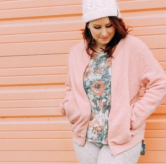 A reviewer wearing pink jacket, colorful pink top and white hat leaning on pink wall