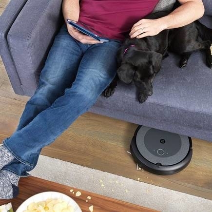 A roomba cleaning up crumbs while the owner relaxes on the couch