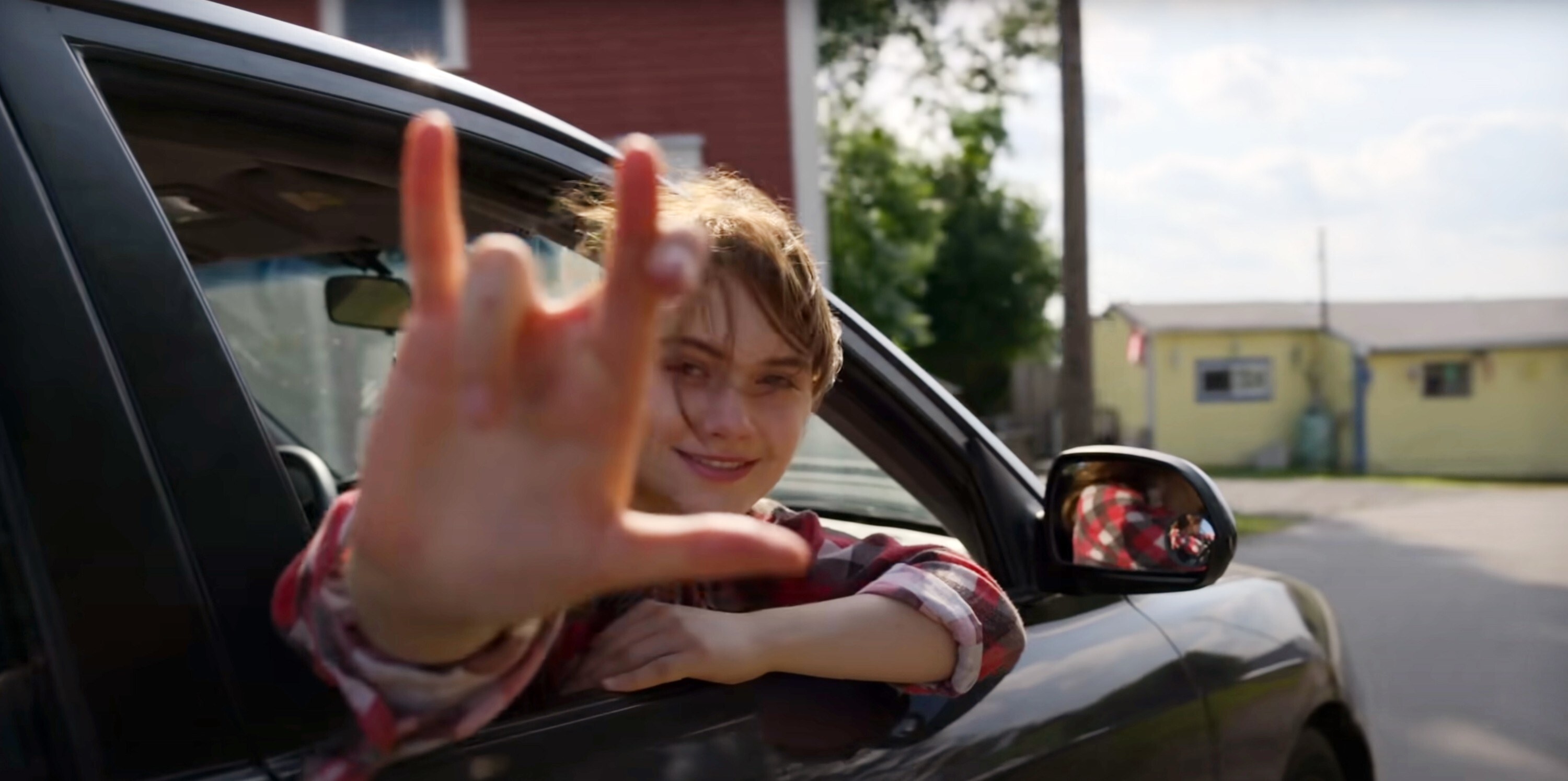 A white girl with brown hair sits in the passengers seat of a car. She looks back out the window signing, "I love you" in sign language