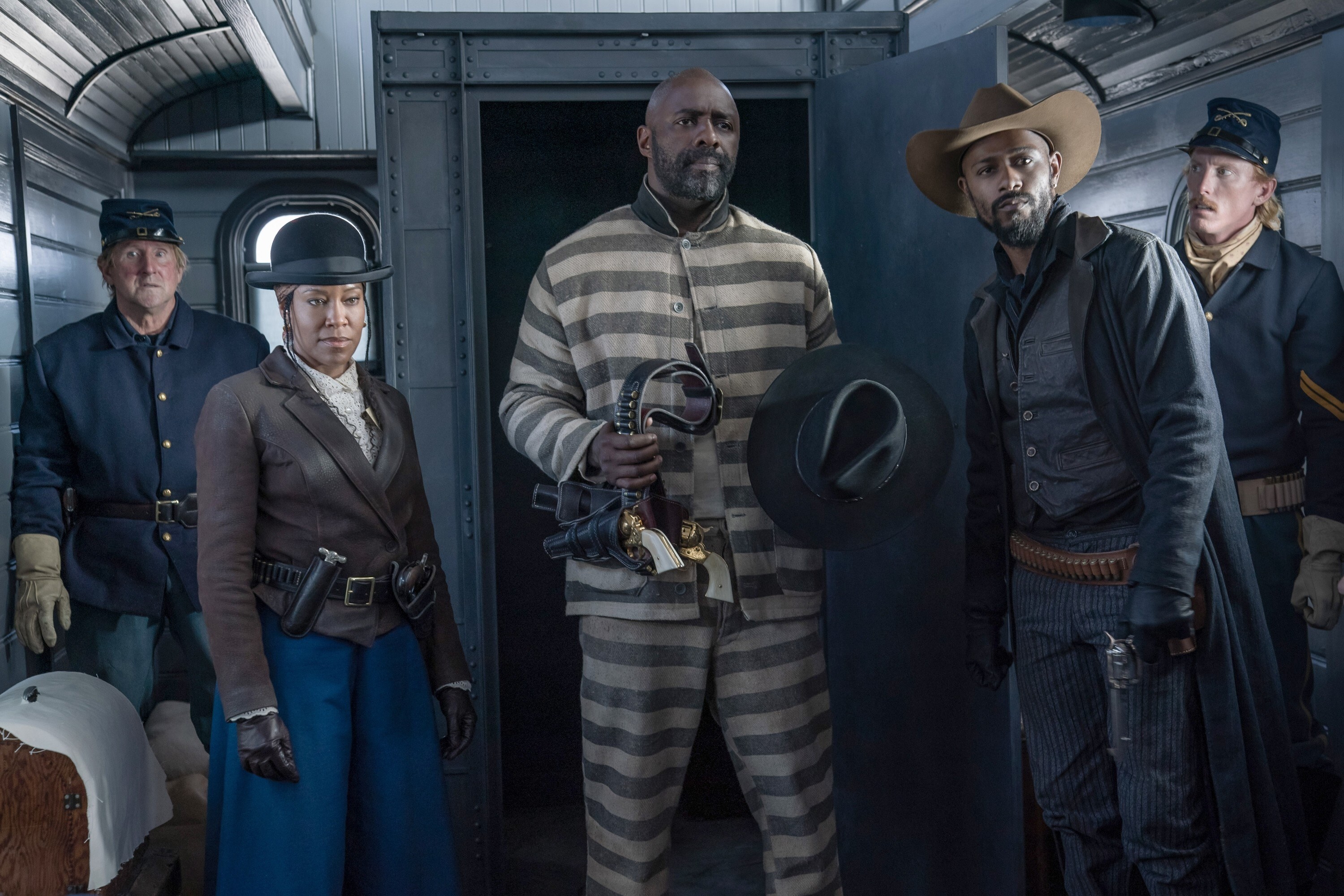 A Black man stands in a prison uniform, he holds a black hat and gun. A Black woman and Black man both in hats and guns stand on either side