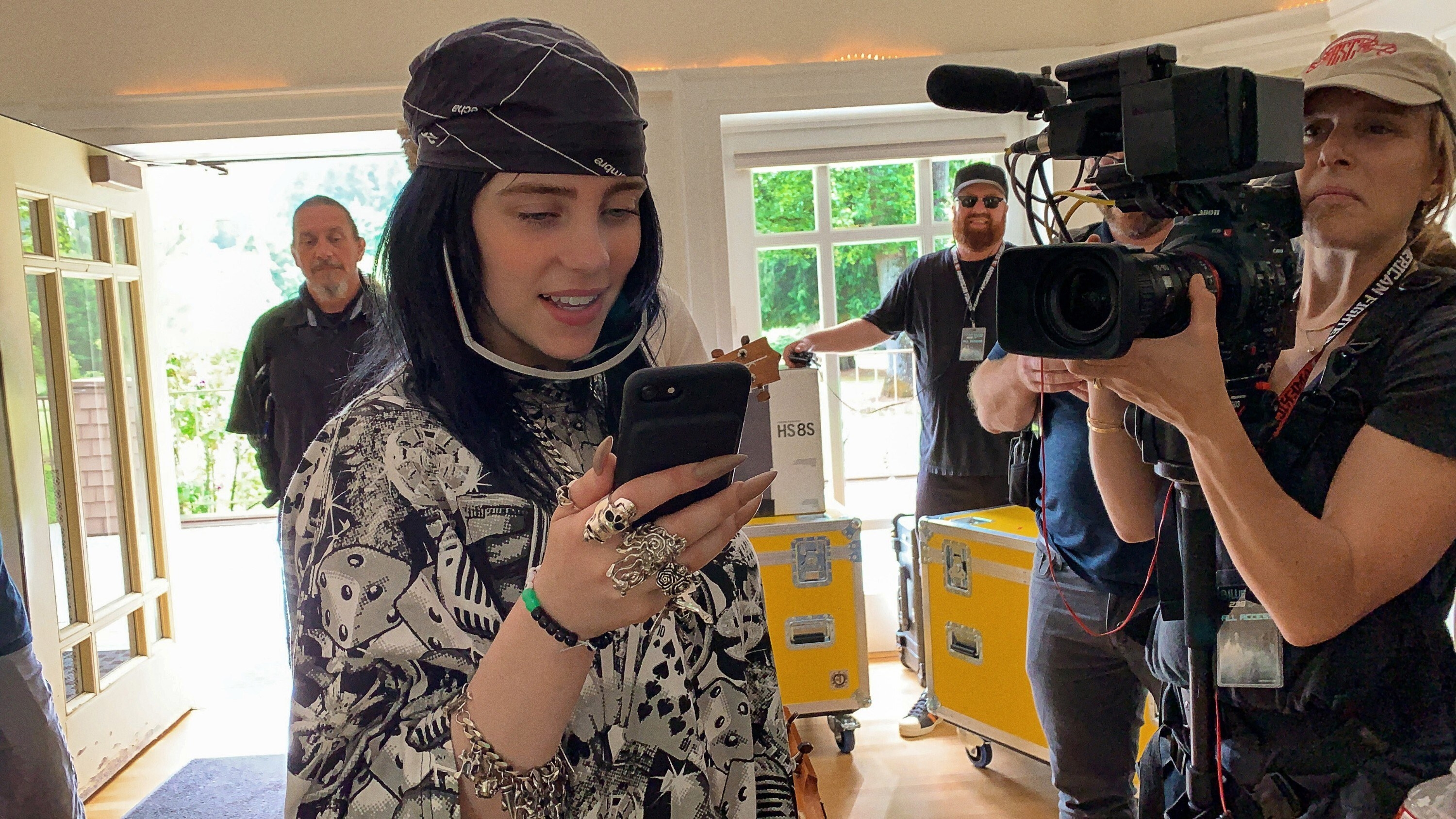 Billie Eilish, a white teenage girl, stands in a room looking at her phone. She wears a bandana in her hair and has many silver rings. A woman holding a movie camera shoots her