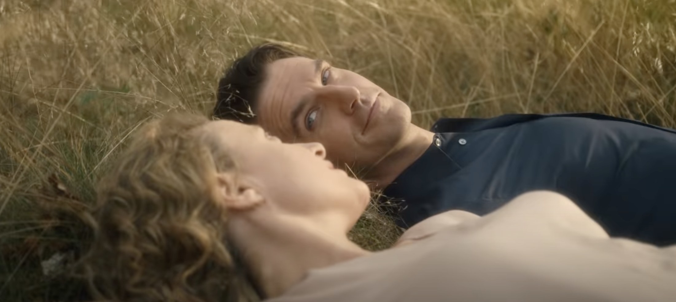 A white man with brown hair and a white woman with blonde hair lie in a field. They look at each other