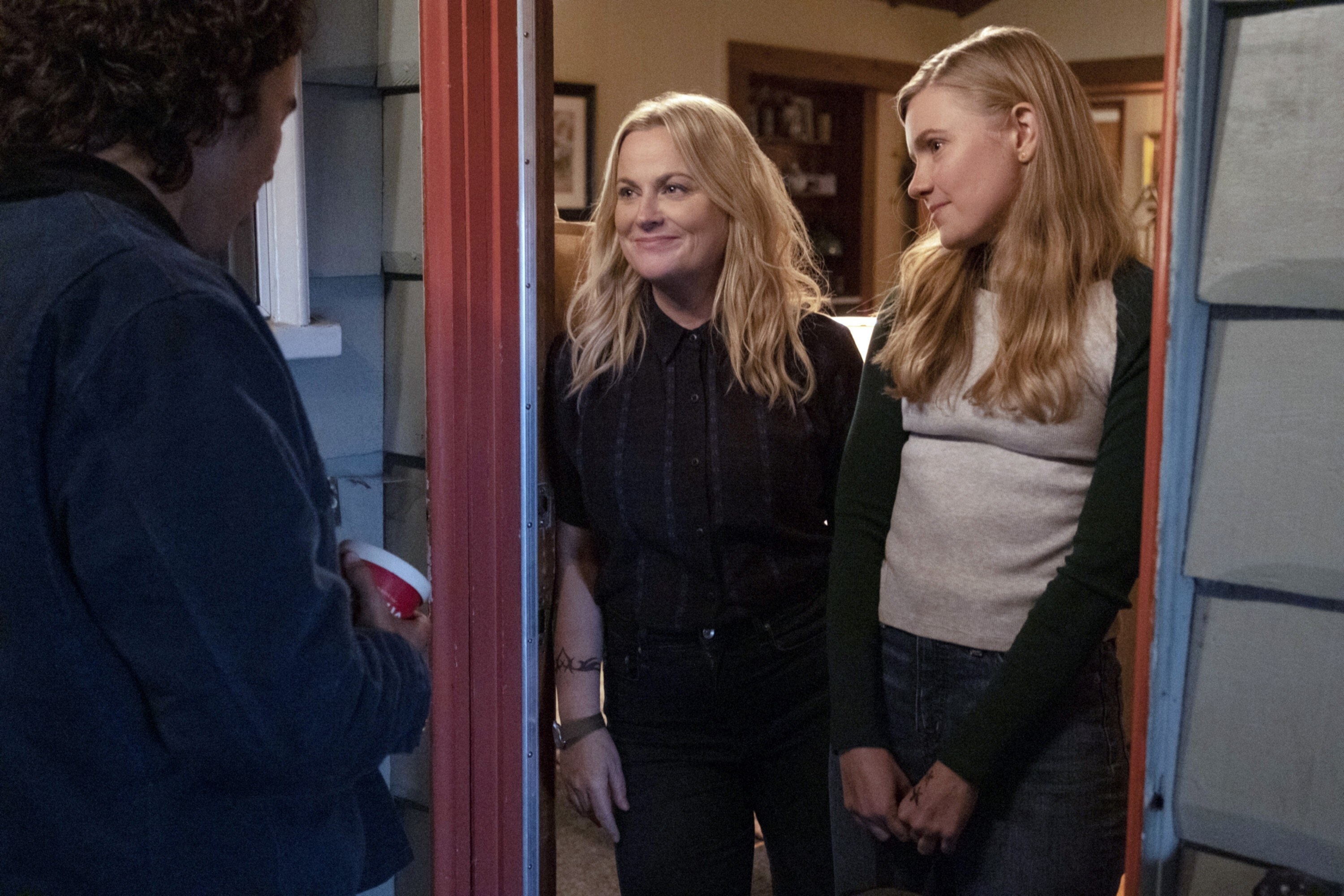 A white mother and daughter, both blonde, stand in the doorway of their home&#x27;s front door, looking at a man with brown hair. They both are smiling