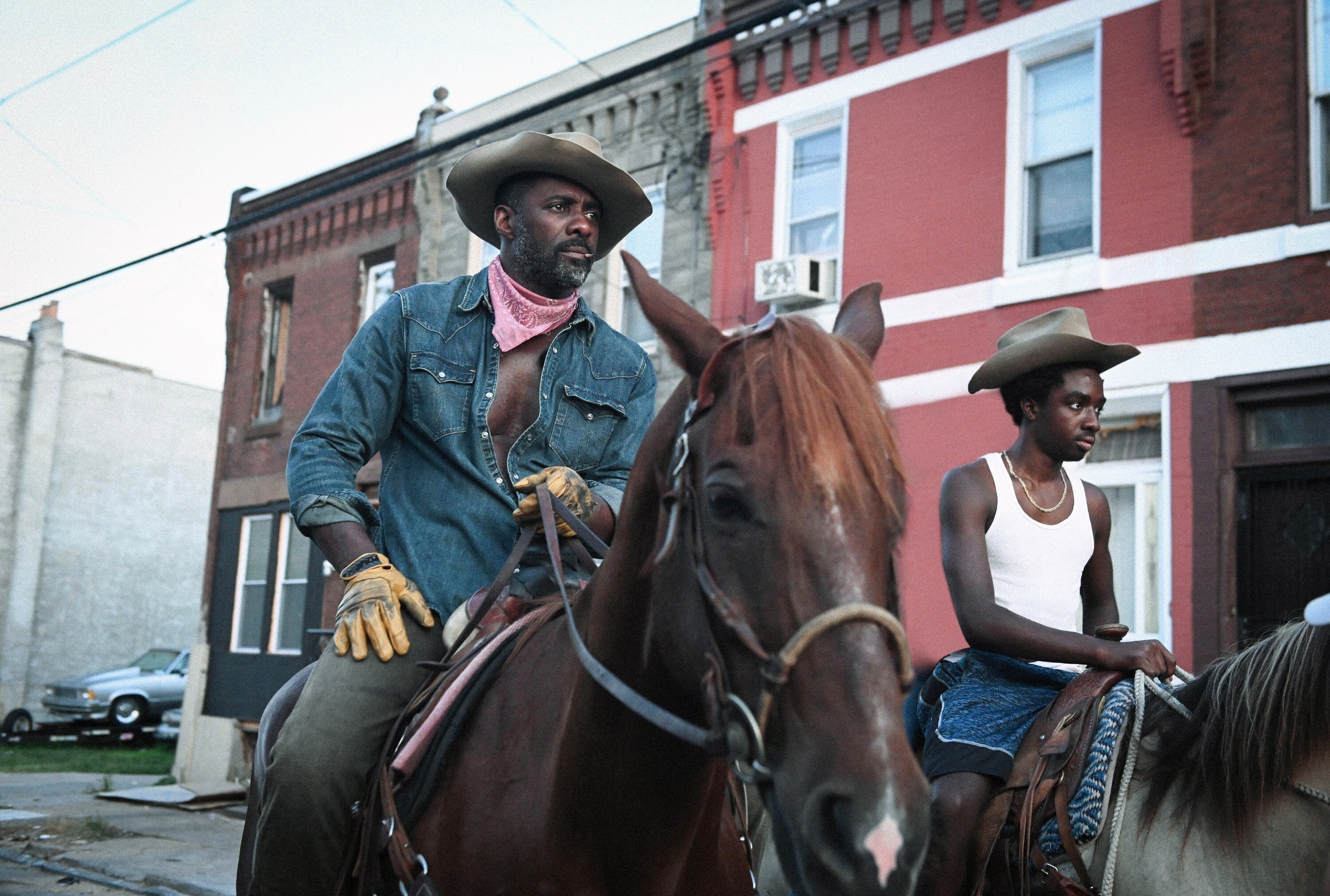 A Black man rides a horse through a street in Philadelphia; he wears a cowboy hat and bandana — and a Black teenage boy rides another horse next to him