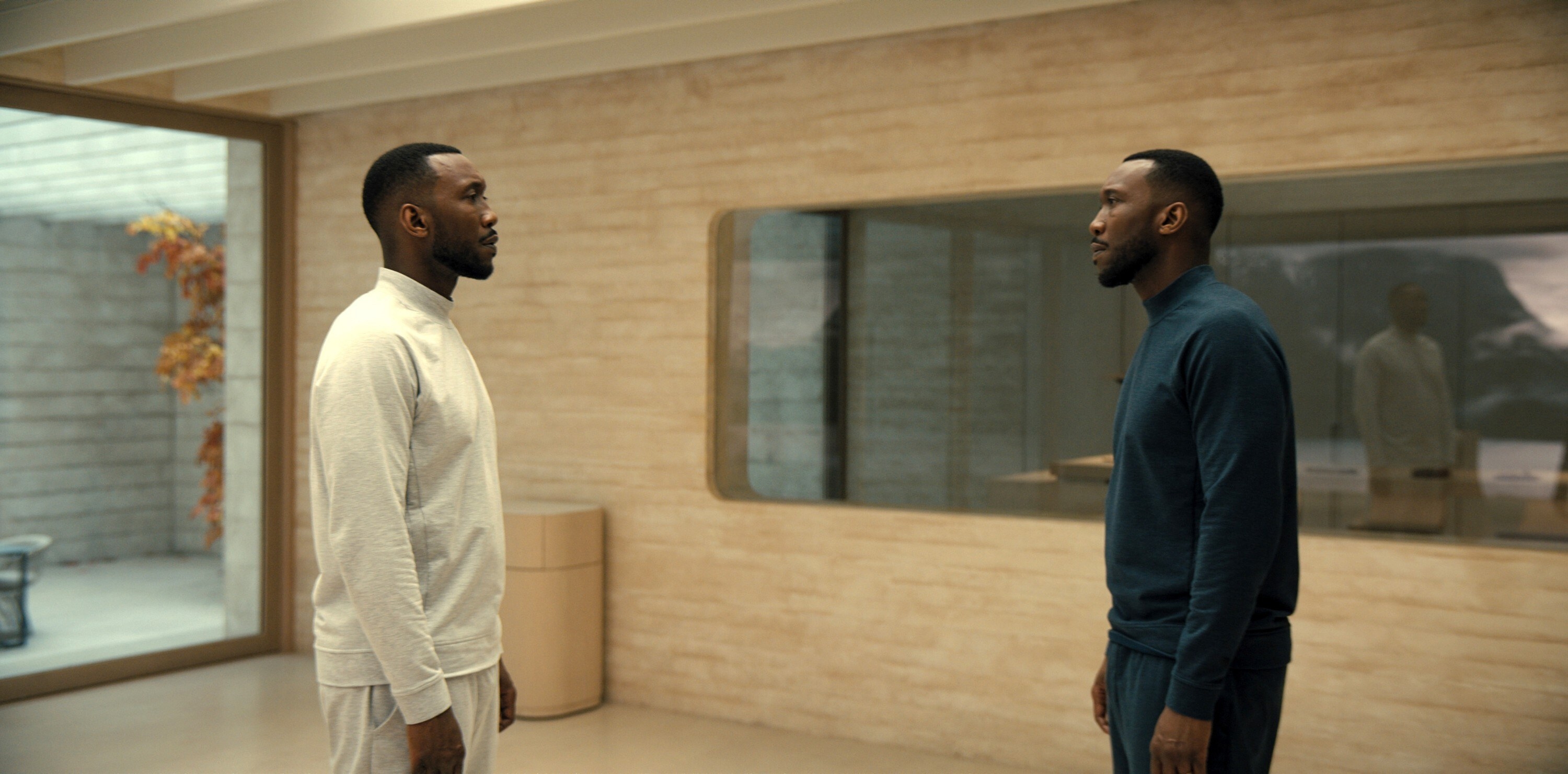 Two identical Black men stand staring at each other in a spare room. One wears white one wears navy blue