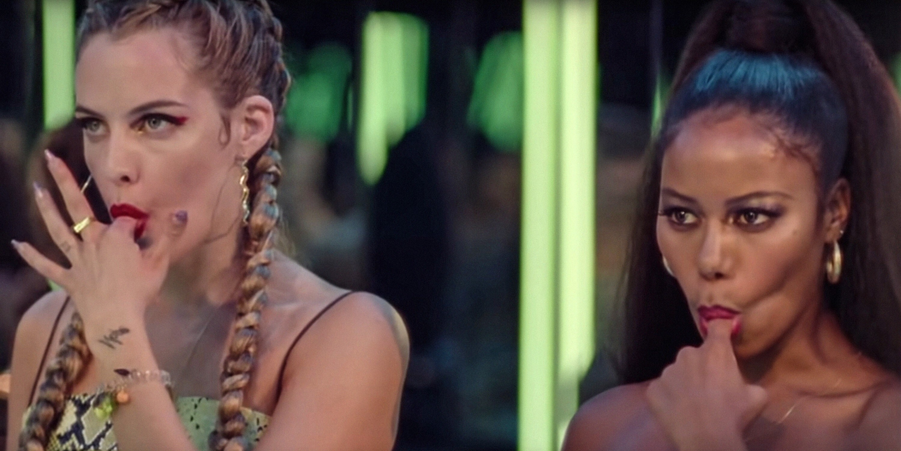 A white girl with two blonde French braids and a Black woman with a high ponytail stand behind bright green lights. They both have their own pointer finger in their mouth