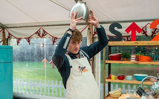 comedia james acaster standing in the bake off tent with a mixing bowl over his head