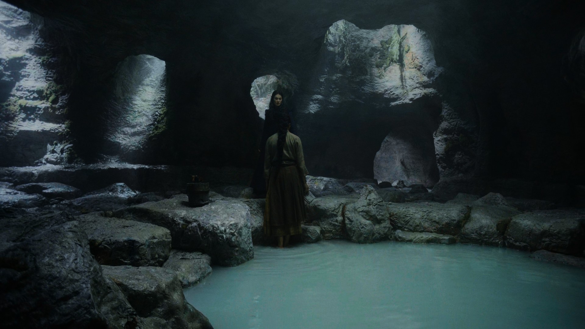 Moiraine and Nynaeve confront each other in a dimly lit cave, with Nynaeve standing in an ankle-deep opaque pool of water
