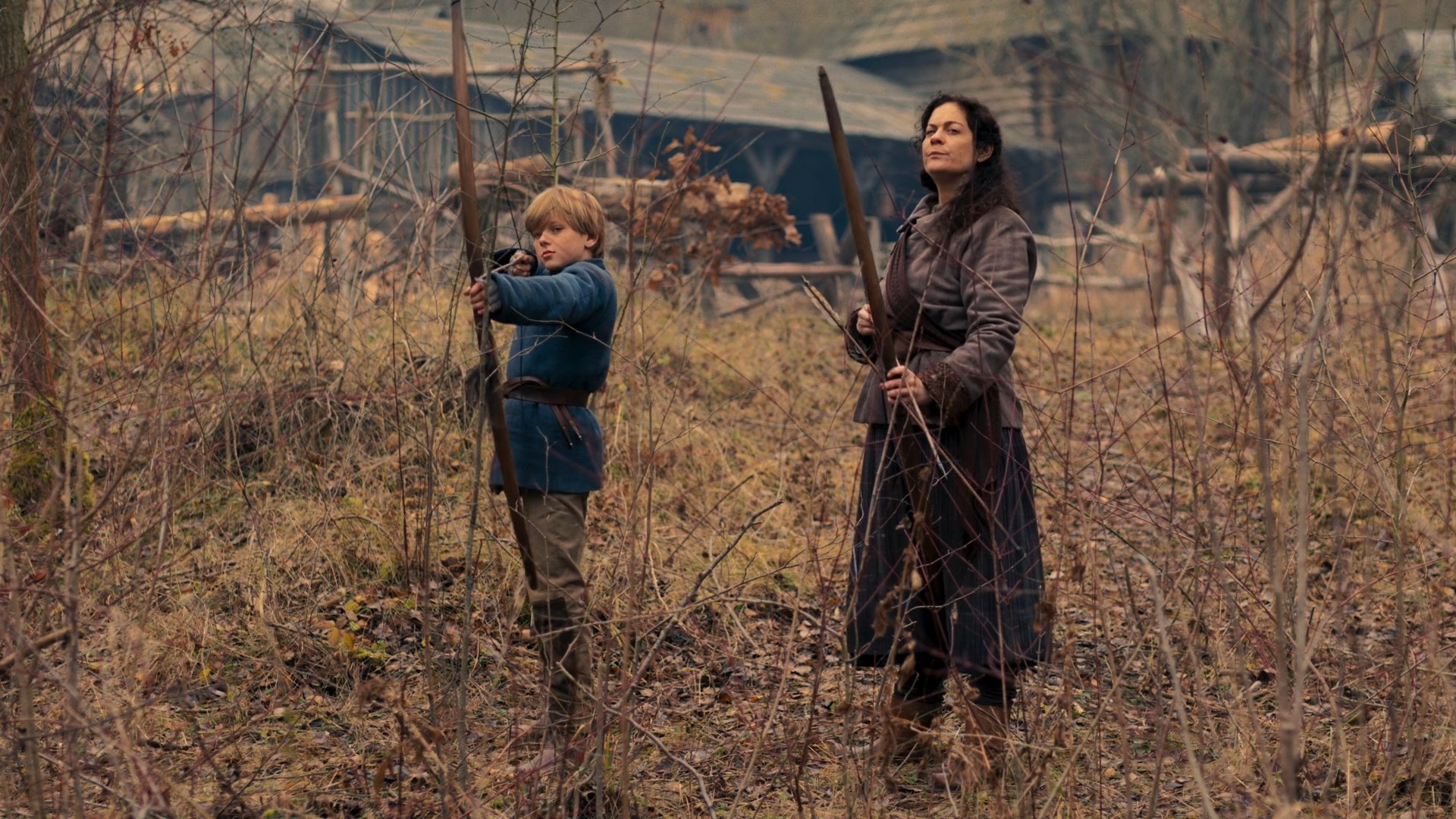 A shot of Mrs Grinwell (Jennifer Preston) and her son (David Dvorscik) with bows drawn outside of a rural farm. They look on suspiciously as they stand in the thicket.