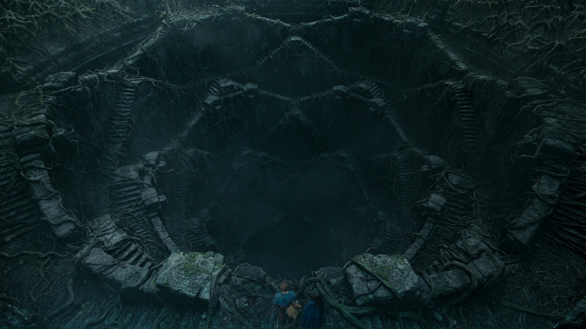 An overhead shot of Moiraine and Rand looking down into a large, open pit, the Eye of the World. The walls of the Eye are lined with triangular staircases leading down into a murky bottom