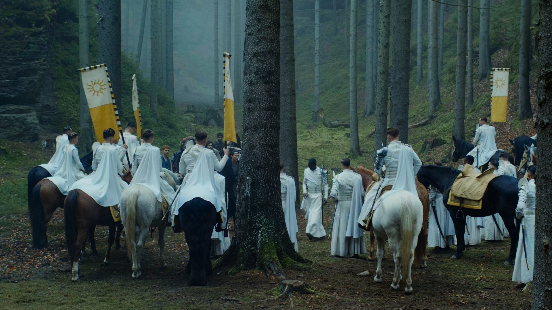 A wide shot of a group of a dozen white-garbed men, some on horses, some on foot, standing in the woods. Moiraine, Lan, Egwene, Mat, Rand and Perrin are facing the men