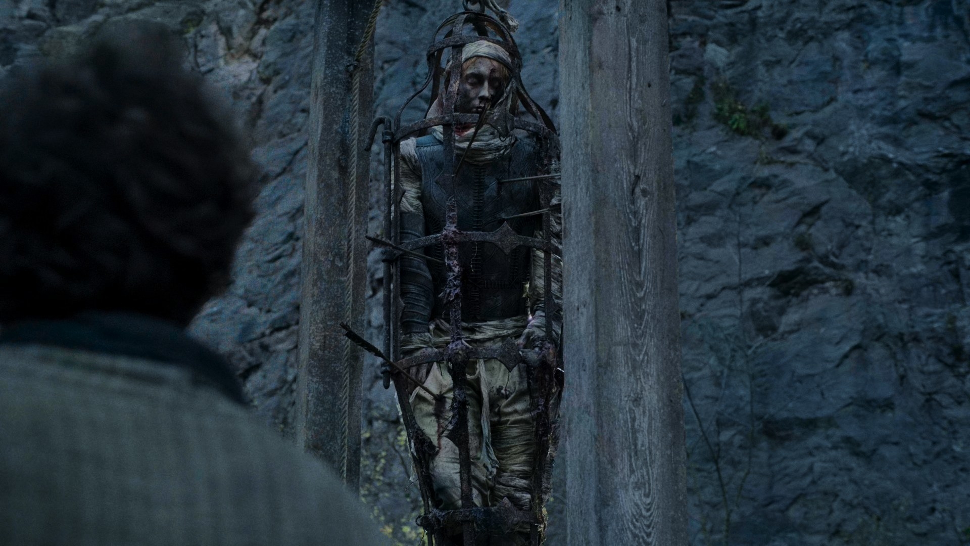 A man in beige clothing and a headscarf is in a cage hanging from a gibbet. He is pierced with many arrows through his torso, which has a stylised leather jerkin on it. He has been dead for a while
