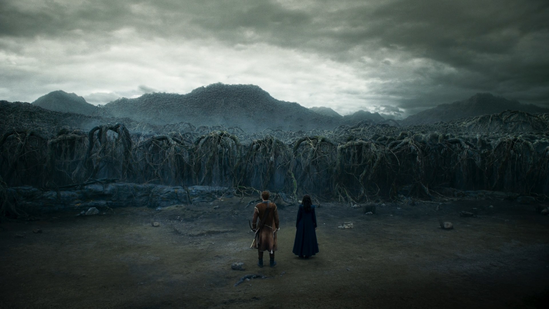 A wide shot of Moiraine and Rand headed towards a gnarled, unwelcoming forest of twisted, diseased trees that stretches on towards the horizon