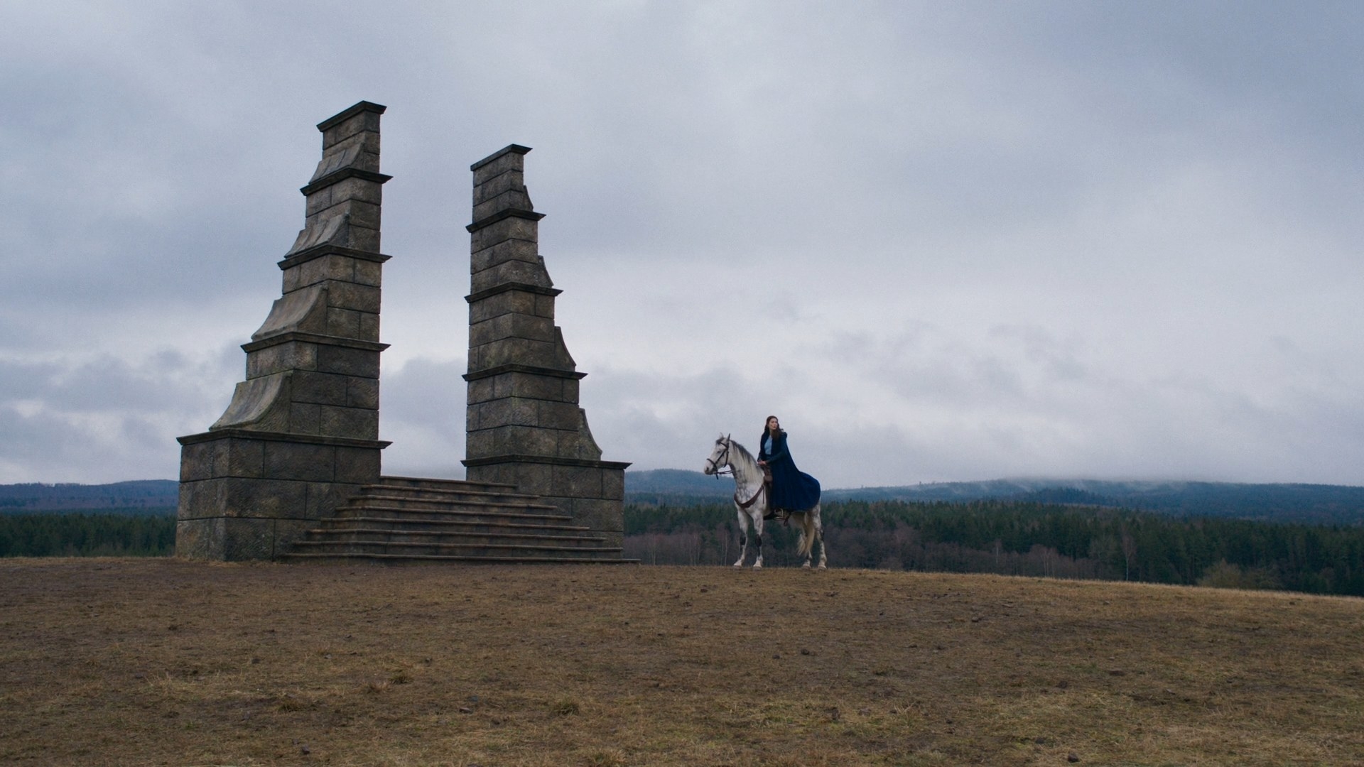 A long shot of Moiraine on her white horse, Aldieb. She is on a treeless hilltop with short brown grass in front of a large stone structure. It has two tall sides rising from a set of steps up towards an opening, but seemingly leads to nowhere
