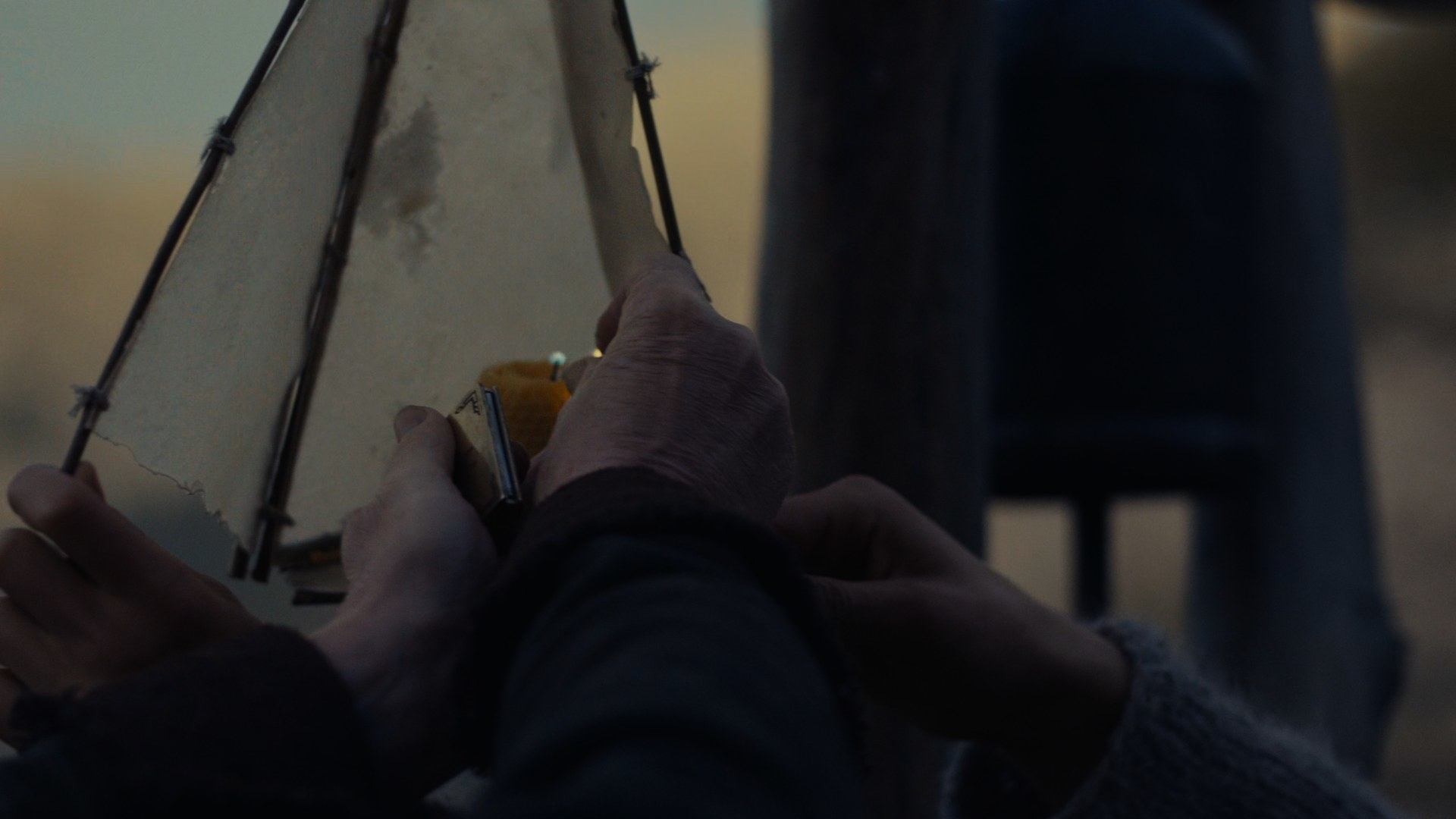 A close-up shot of Tam (Michael McElhatton) and Rand’s (Josha Stradowski) hands as they light a candle within a paper lantern. In Tam’s left hand is a shiny metal book of matches.