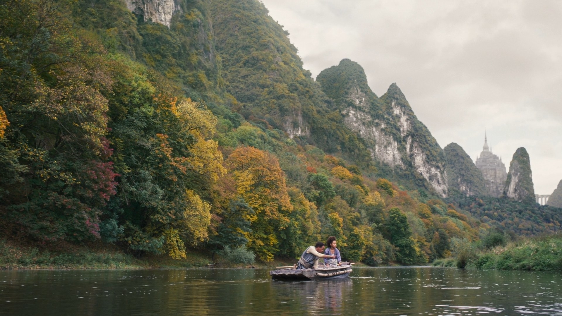 A shot of young Siuan and her father in a fishing skiff on the river. In the background, nearly blending into the mountain peaks, is what appears to be a large fortress.