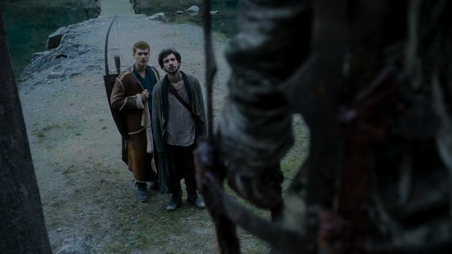 A shot of Rand and Mat as they look upon the caged dead man. Rand, in a long sheepskin coat and carrying an arrow and quiver, is muttering something to Mat, who is wearing colourless, threadbare clothes.