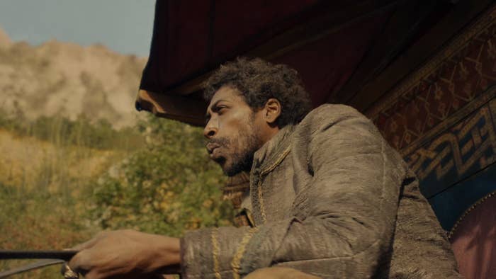 A low-angle shot of a bearded Padan Fain (Johann Myers), in a brown coat, holding the reins to a team of horses and with his lips pursed, as if whistling