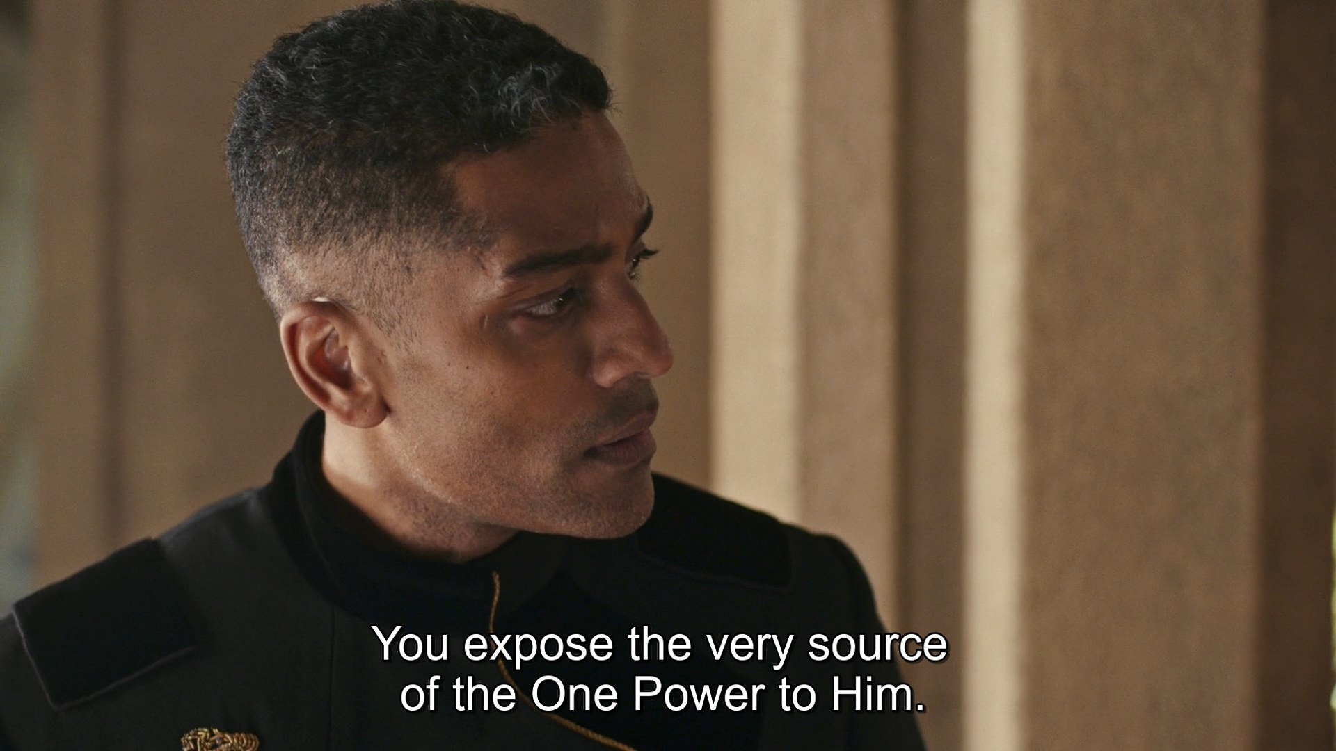 A close-up shot of Lews Therin Telamon (Alexander Karim), the Dragon in the Age of Legends, in a black uniform. The subtitles, translated from the Old Tongue, reads “You expose the very source of the One Power to Him”, words spoken by Latra.