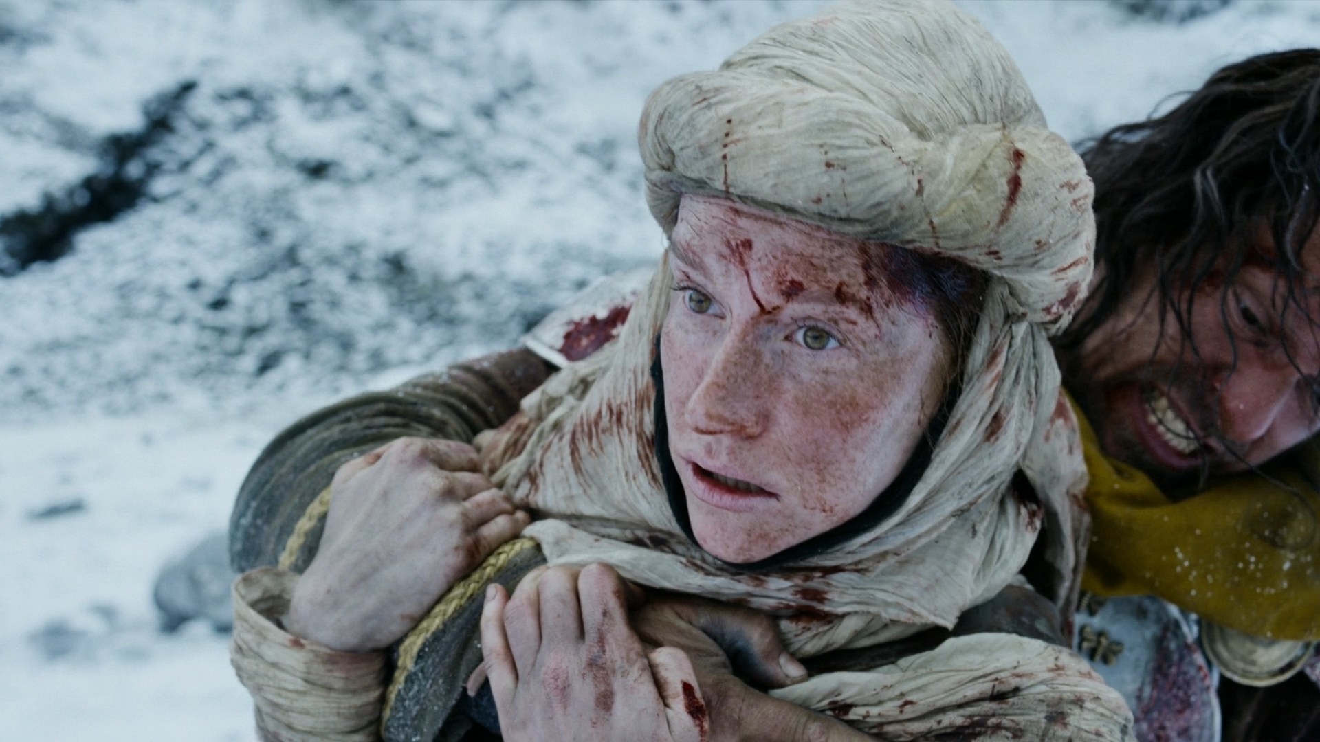 A close-up shot of an Aiel woman (Magdalena SIttova) with a white-beige shoufa wrapped around her head. She is spattered in blood, and a man in a yellow cape is holding her from behind. Her face is a mask of shock.
