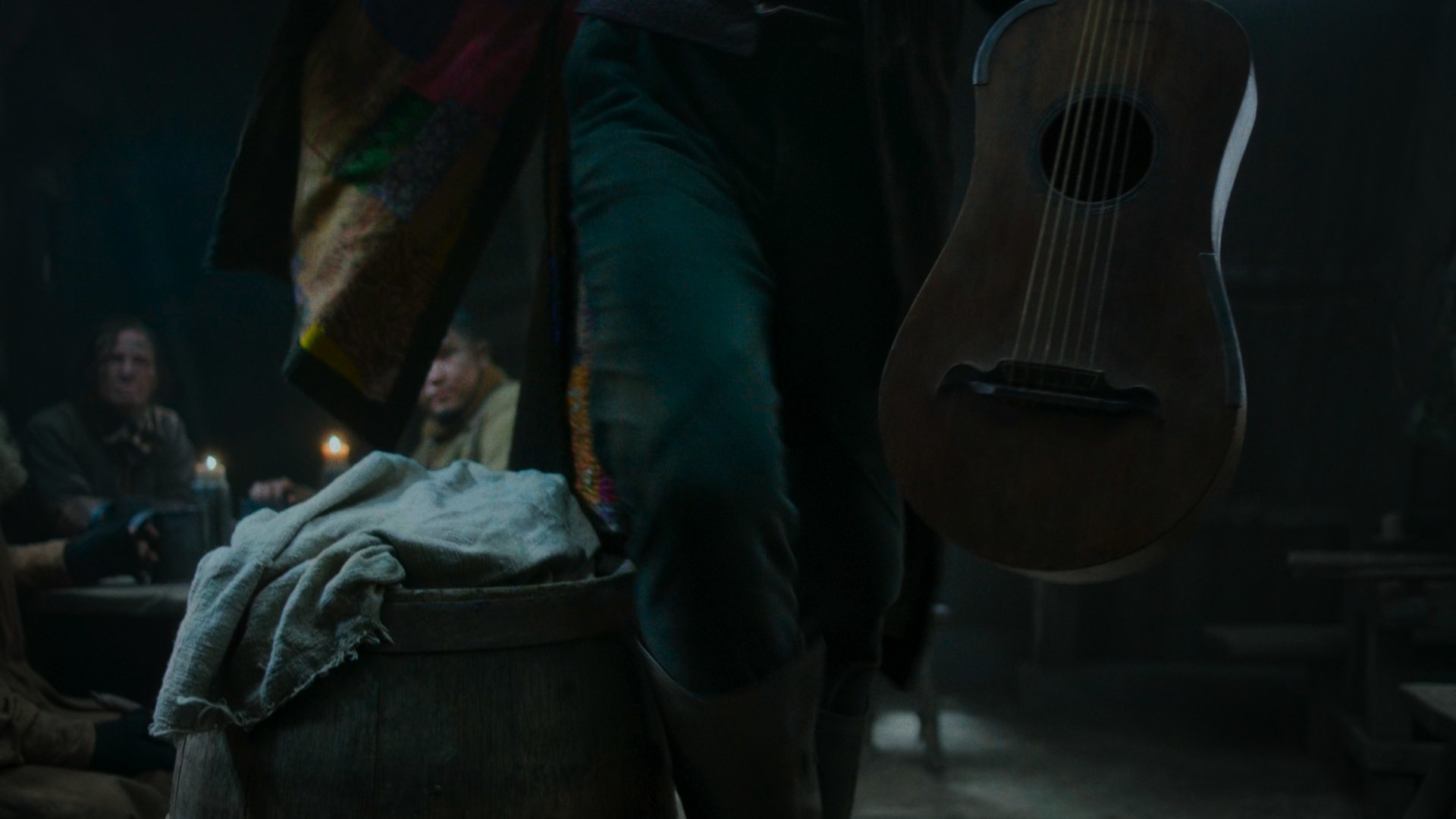 A shot of Thom Merrilin’s (Alexandre Willaume) lower body as he picks up a wooden guitar and sits on a propped-up barrel. On the inside of his coat are a number of colourful patches.